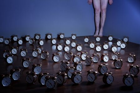 Bare legs, feet  and hands seen against a blue wall in a room where the brown floor is covered by alarm clocks. の写真素材
