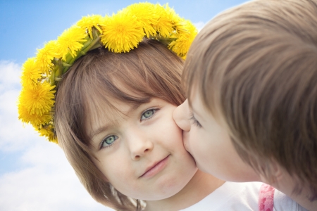 Young boy kissing cute preschool sister with yellow daisy flower crown, blue sky and cloudscape background.の写真素材
