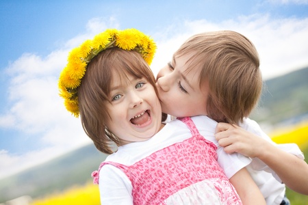 Young boy kissing cute preschool sister with yellow daisy flower crown, blue sky and cloudscape background.の写真素材