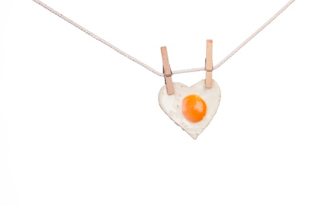 Fried egg in shape of love heart hung from wooden clothes pins or pegs, white background の写真素材