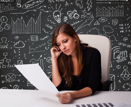 Pretty young businesswoman sitting at desk with business scheme and iconsの写真素材