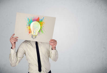 Businessman holding a cardboard with coloured paint splashes and lightbulb in front of his headの写真素材