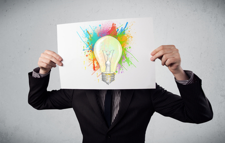 Businessman holding a cardboard with coloured paint splashes and lightbulb in front of his headの写真素材