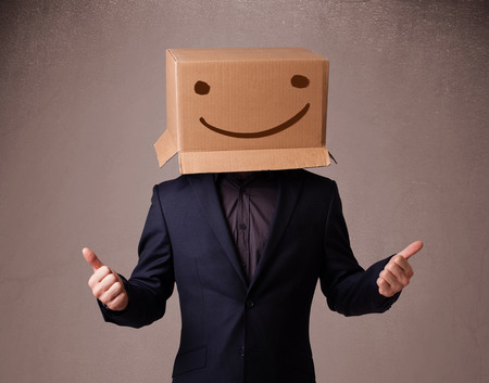 Young man standing and gesturing with a cardboard box on his head with smiley faceの写真素材
