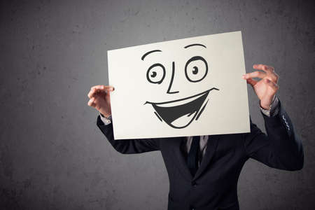 Young businessman holding a cardboard with a smiley face on it in front of his headの写真素材