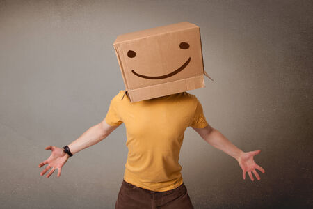Young man standing and gesturing with a cardboard box on his head with smiley faceの写真素材