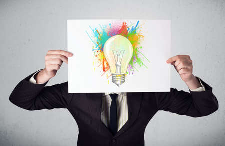 Businessman holding a cardboard with coloured paint splashes and lightbulb in front of his headの写真素材
