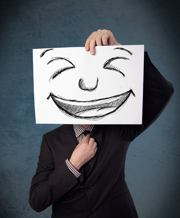 Businessman holding a paper with a drawed smiley face on it in front of his headの写真素材