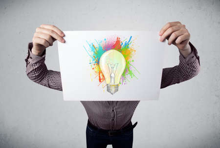 Businessman holding a cardboard with coloured paint splashes and lightbulb in front of his headの写真素材