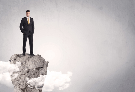 A happy successful businessman standing on a stone cliff with clouds in front of clear empty grey background conceptの写真素材