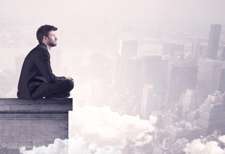 An elegant young businessman sitting on the edge looking over urban city landscape with clouds conceptの写真素材