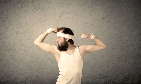A young man with beard, headstrap and glasses posing in front of blank grey wall background, imagining he has big musclesの写真素材