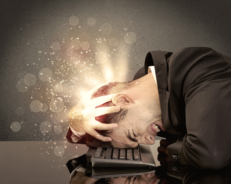 A young depressed business person laying his head on computer keyboard with thoughts exploding from his head illustrated by light beams conceptの写真素材