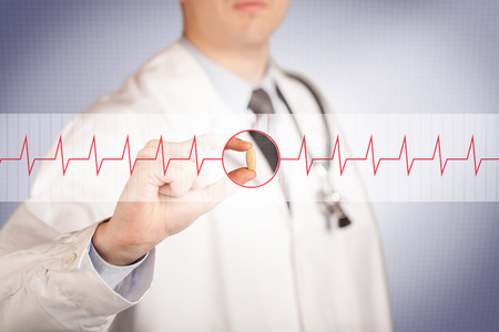 A male doctor in white coat with a stethoscope on one shoulder holding a pill between his fingers focused on a heartbeat graphの写真素材