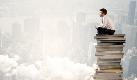 A serious businessman in elegant suit sitting on a pile of giant books in front of a grey city scape with clouds, fogの写真素材