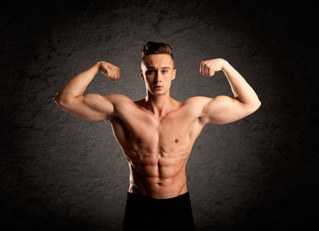 A handsome fitness guy lifting weight and showing his strong muscles in front of a black urban concrete wall conceptの写真素材