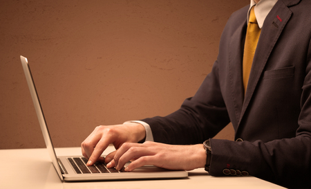 An office worker in elegant suit sitting at desk, typing on portable laptop with empty brown wall backgroundの写真素材