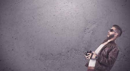 A stylish funny hipster person holding a vintage camera and taking photographs in front of a concrete clear empty urban wall background conceptの写真素材