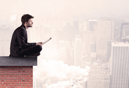 An elegant businessman in modern suit sitting on the top of a brick building, looking over the cityscape with clouds conceptの写真素材