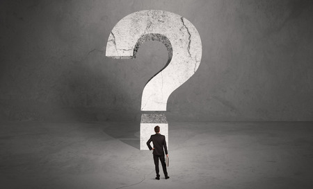A small elegant business person in suit standing with his back in front of a huge question mark in open space conceptの写真素材