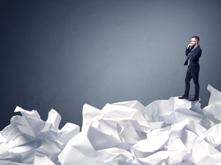 Thoughtful young businessman standing on a pile of crumpled paper with a blueish grey backgroundの写真素材