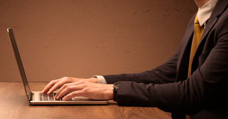 An office worker in elegant suit sitting at desk, typing on portable laptop with empty brown wall backgroundの写真素材