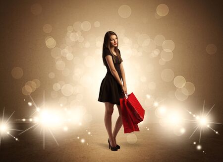 A beautiful elegant woman in black standing with red shopping bags in front of brown background and bright glowing lights conceptの写真素材