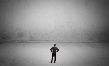 A tiny elegant businessman standing in large empty urban space with concrete walls and grey background conceptの写真素材
