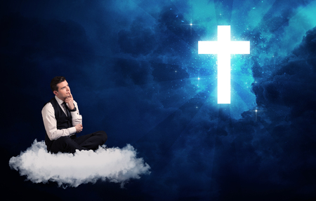 Caucasian businessman sitting on a white fluffy cloud looking and wondering at a big bright glowing crossの写真素材