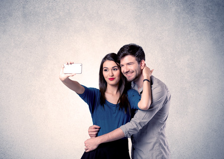 A young couple in love taking selfie with a mobile phone in the handsome guy's hand in front of an empty clear grey wall background conceptの写真素材