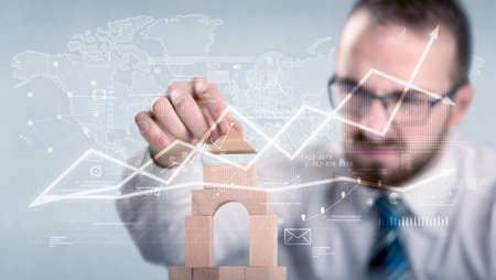 Young handsome businessman using wooden building blocks with data and calculations behind himの写真素材