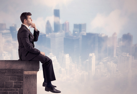 A young businessman sitting at the edge of a building in front of a city center scape background with tall buildings and clouds conceptの写真素材
