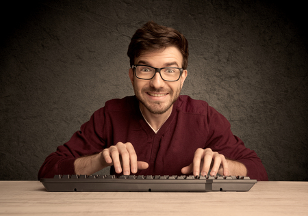 A young hacker with glasses dressed in casual clothes sitting at a desk and working on a computer...