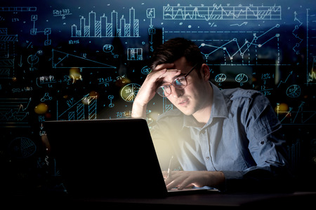 Young handsome businessman working late at night in the office with blue calculations in the...