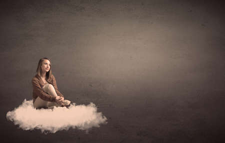Caucasian woman sitting on a white fluffy cloud daydreaming beside a plain grunge backgroundの写真素材