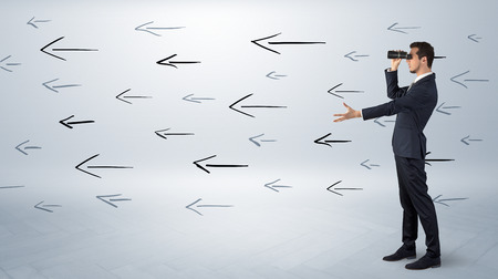 Young businessman standing and looking through binoculars with drawn arrows aroundの写真素材