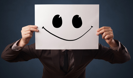 Casual person holding a paper with funny emoticon in front of her faceの写真素材