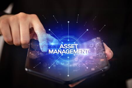 Businessman holding a foldable smartphone with ASSET MANAGEMENT inscription, new business conceptの写真素材