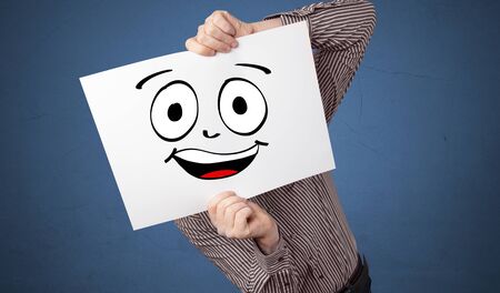 Young student holding a paper with laughing emoticon in front of his faceの写真素材