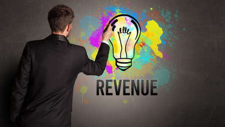 businessman drawing colorful light bulb with REVENUE inscription on textured concrete wall, new business idea conceptの写真素材