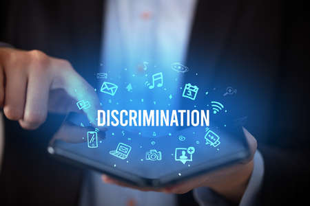 Businessman holding a foldable smartphone with DISCRIMINATION inscription, social media conceptの写真素材