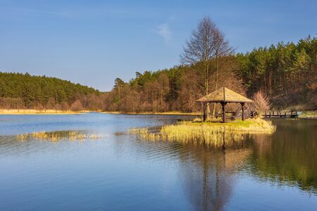 Small lake surrounded by forest. Wooden gazebo and bridge on the water. Dywan, Pomerania, Poland.の写真素材