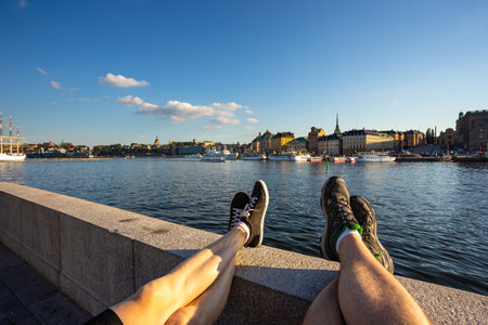 Stockholm, Sweden - 25 Juny 2016: Men and women legs in black sports shoes and navy blue jeans on the wall. Old Town, Gamla Stan in the background.のeditorial素材