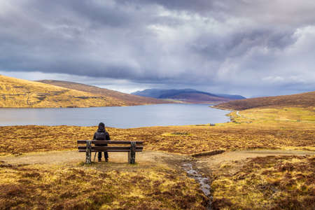 Woman on a bench looking at Leitisvatn Lake. The old path to the peat field, a Traelanipan Trail. Cloudy day on the Vagar island. Faroe Islands.の写真素材