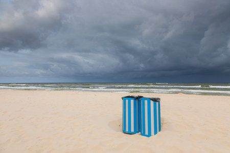 Two blue striped trash bins on a deserted beach in Karwia, Poland, with dramatic storm clouds over the Baltic Sea.の写真素材