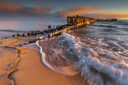 Sunrise light on icy wooden breakwater and beach waves in Babie Doly, Gdynia, Poland. Baltic Sea winter scene.の写真素材