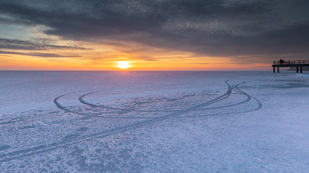 Sunset over the frozen Baltic Sea in Chalupy, Poland, with vehicle tracks on ice and silhouettes on the pier.の写真素材