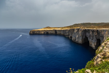 A small boat leaves a white trail on the blue Mediterranean Sea near the limestone cliffs of Gozo Malta.の写真素材