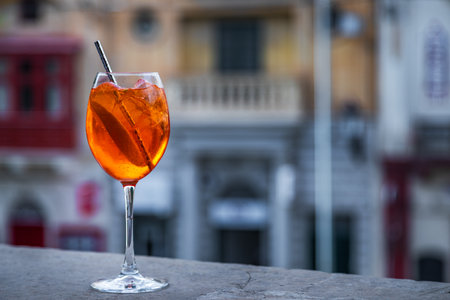 Glass of refreshing Aperol Spritz cocktail with ice cubes, orange slice and straw on a stone ledge, with blurred Mediterranean architecture in the background.の写真素材