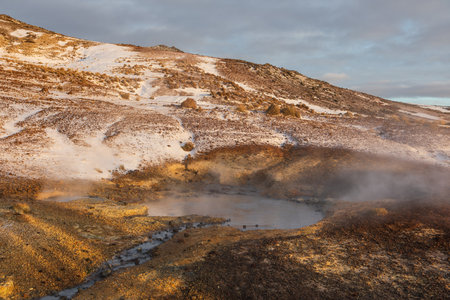 Snow covered geothermal hills rise above shallow pools at Seltun Krysuvik in Iceland. Warm light highlights the mixed volcanic and snowy terrain.の写真素材